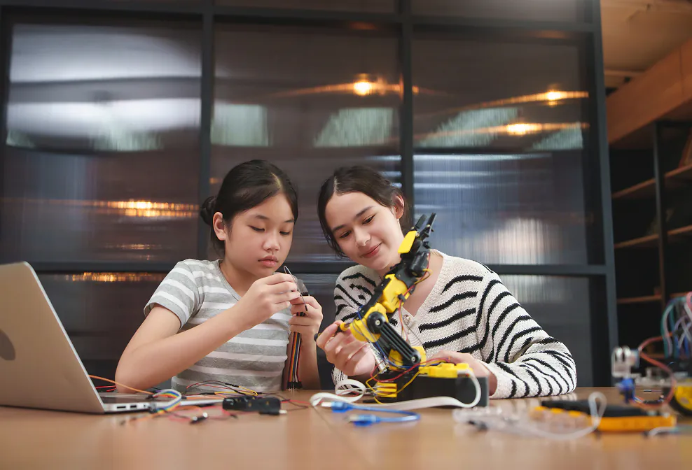 Two young women collaborate on a robotics project, connecting wires and examining a yellow robotic